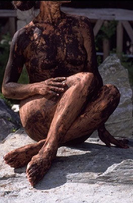 Person with a full-body Moor mud treatment sitting on a rock in a natural outdoor setting.