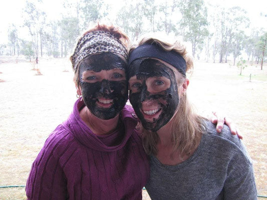 Two women smiling with dark mud masks on their faces, highlighting the fun and social aspect of using the products.