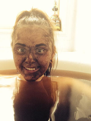A young woman smiling brightly in a white bathtub, with Moor mud on her face and in the water.
