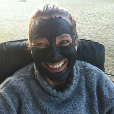 A smiling woman wearing a dark mud mask, showing the fun and effective results of a natural detox treatment.