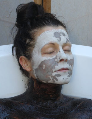 A serene woman with her eyes closed, wearing clay mask on her face and immersed in a warm Moor mud bath.