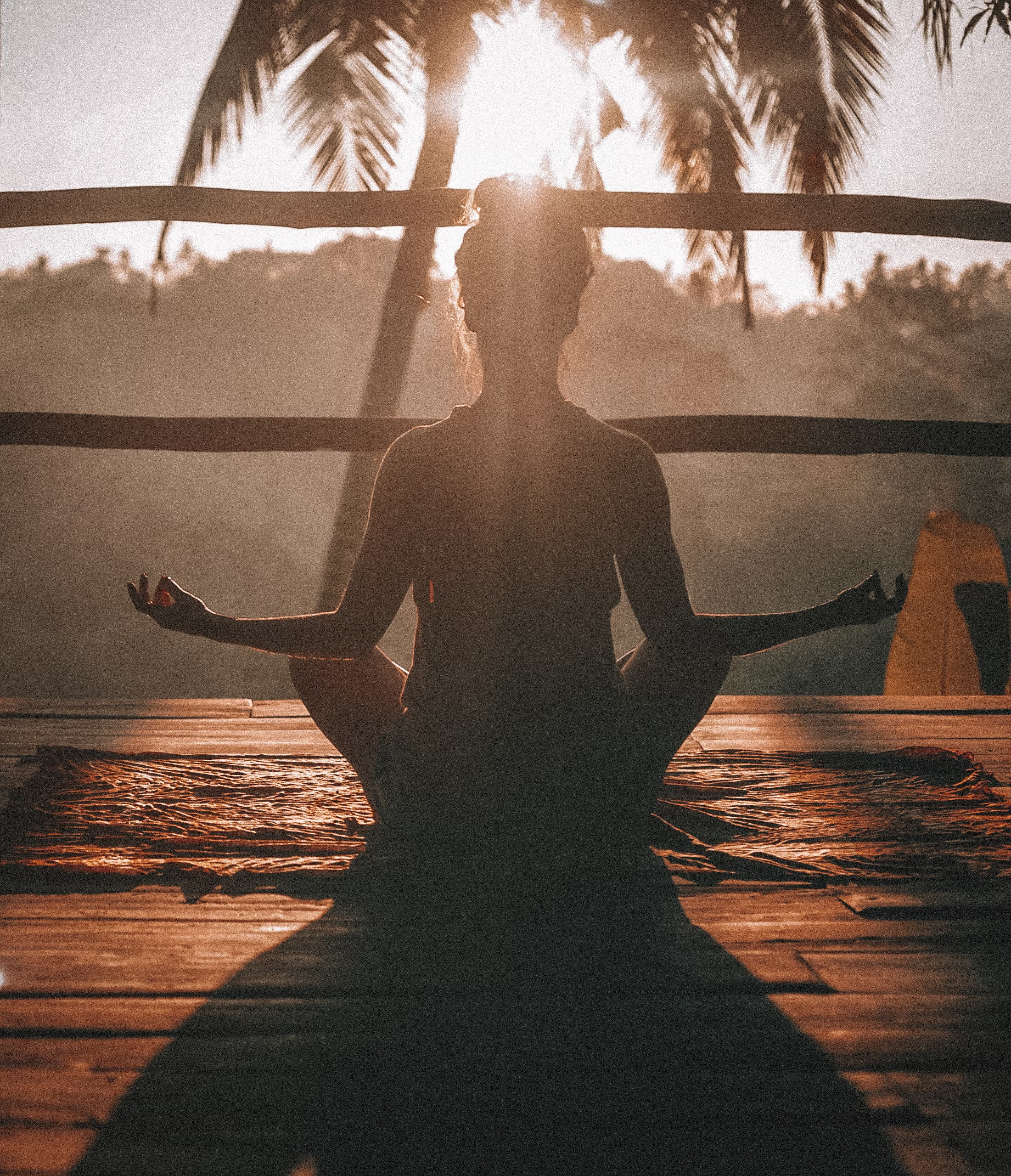A person is in a yoga meditation pose on a wooden deck as the sun rises in the background.